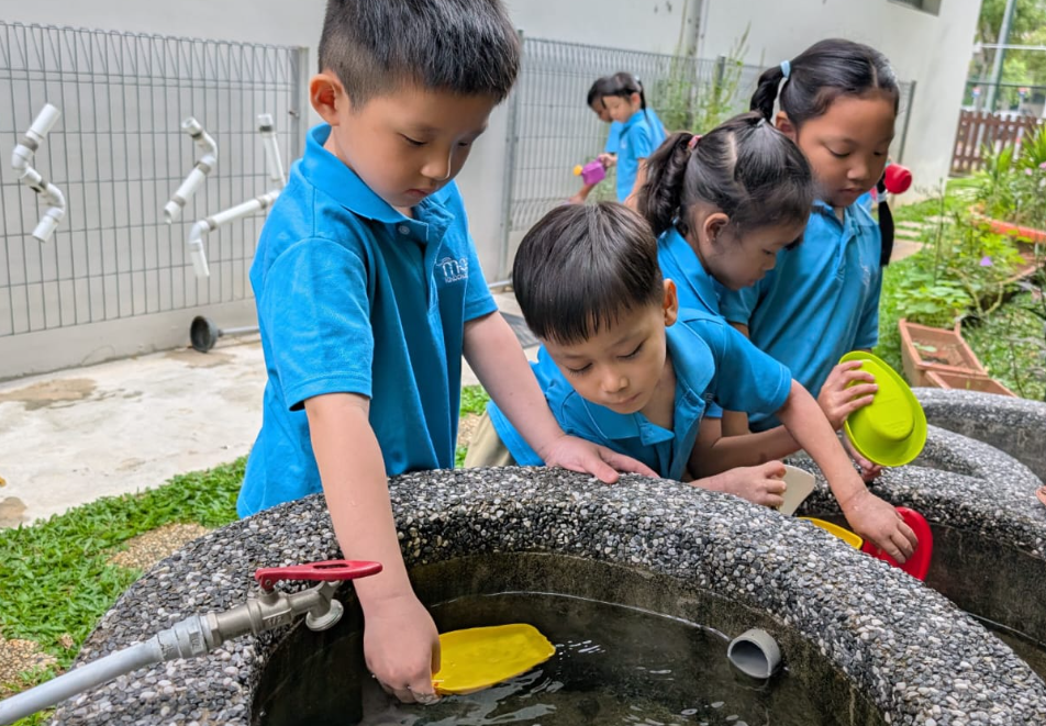 Four children in blue shirts play at an outdoor water basin, dipping colorful plastic bowls in water.